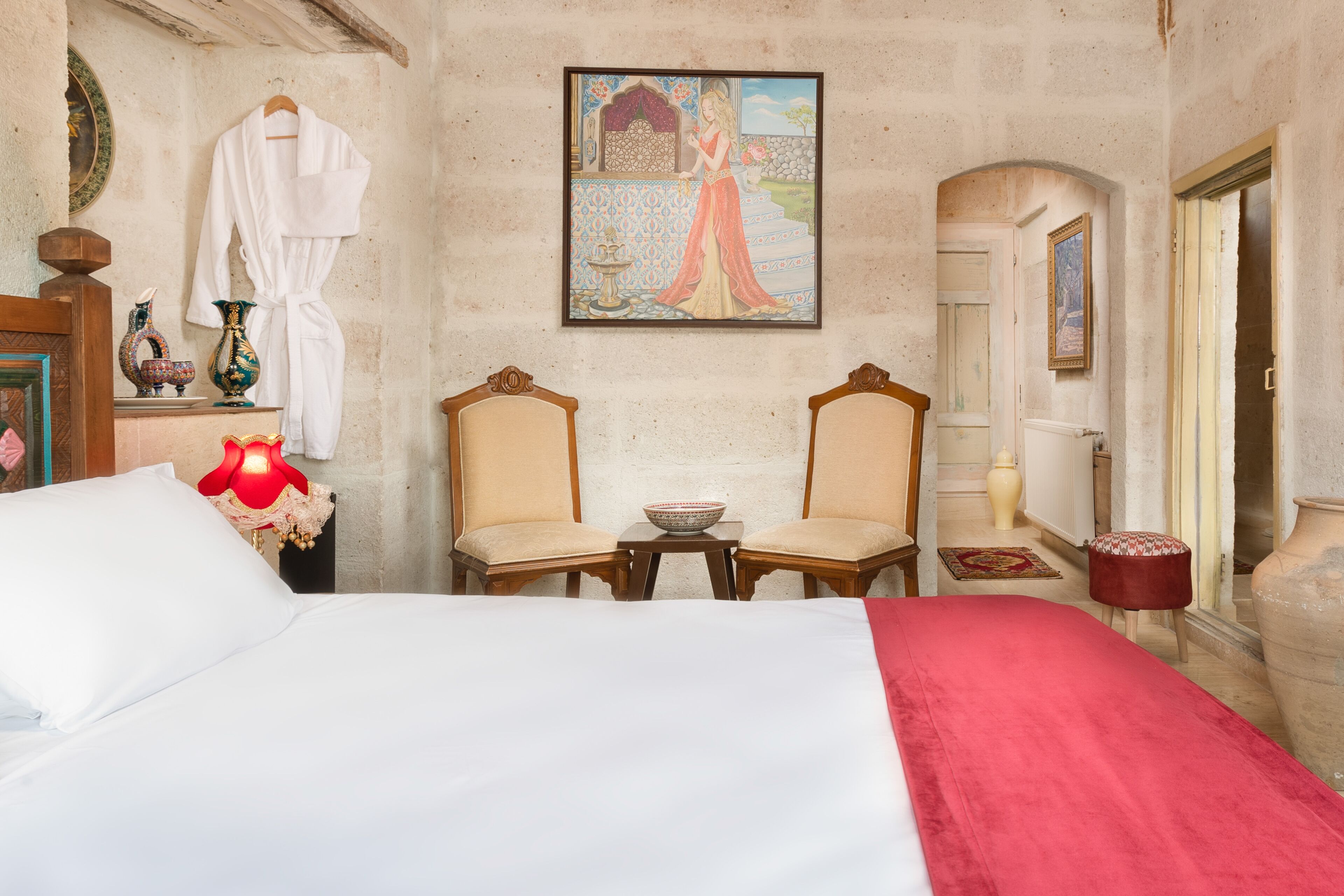 A bedroom with a large bed, two chairs, a small table, a painting, and a vase.
