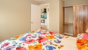 A bedroom with a floral bedspread, a wardrobe, and a view into the kitchen. - Roundham Cottage (Bridport)