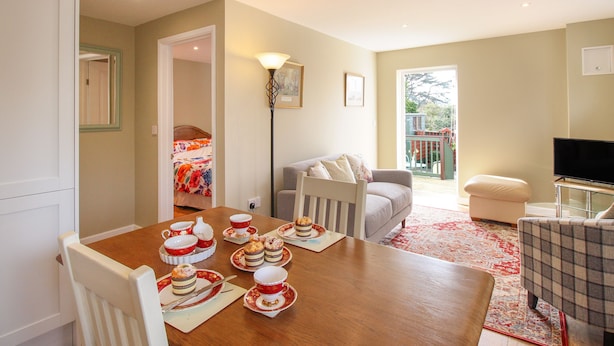 A dining area with a wooden table set for a meal, a sofa, a television, and a view of the outdoors. - Roundham Cottage (Bridport)