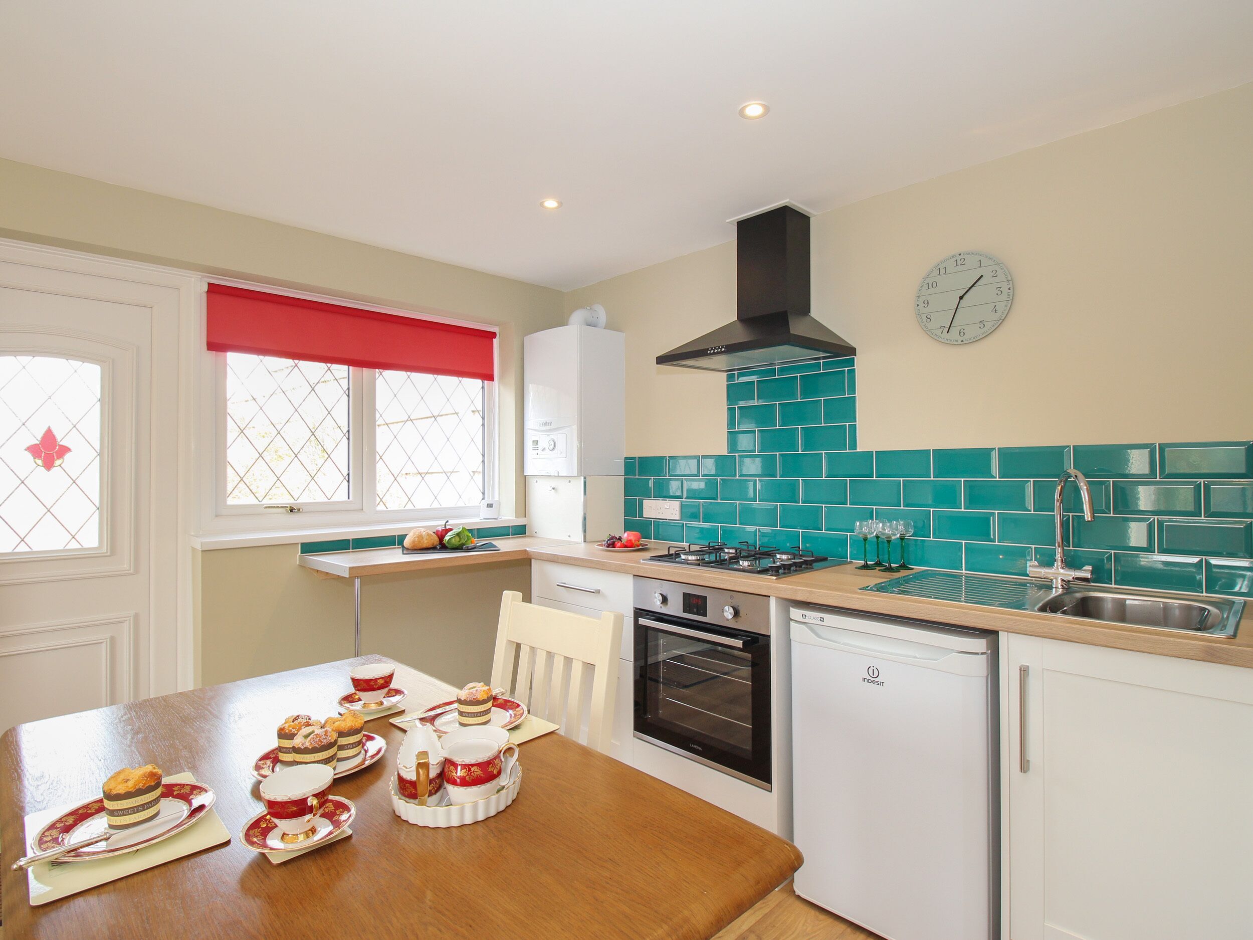 A compact kitchen with a stove, oven, and sink, a dining table with tea sets, and a window with a red valance.
