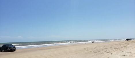 A beach with a truck parked on the sand, a clear sky, and the ocean in the background.