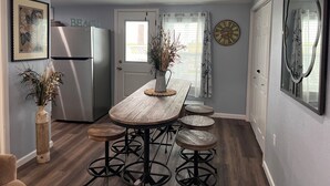 Una cocina con una mesa de madera, taburetes de bar, ventilador de techo, refrigerador y un reloj en la pared.