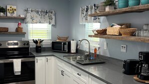 Una cocina moderna con armarios blancos, horno de acero inoxidable, microondas y cafetera. Hay estantes con objetos decorativos y un letrero que dice 'KITCHEN'.