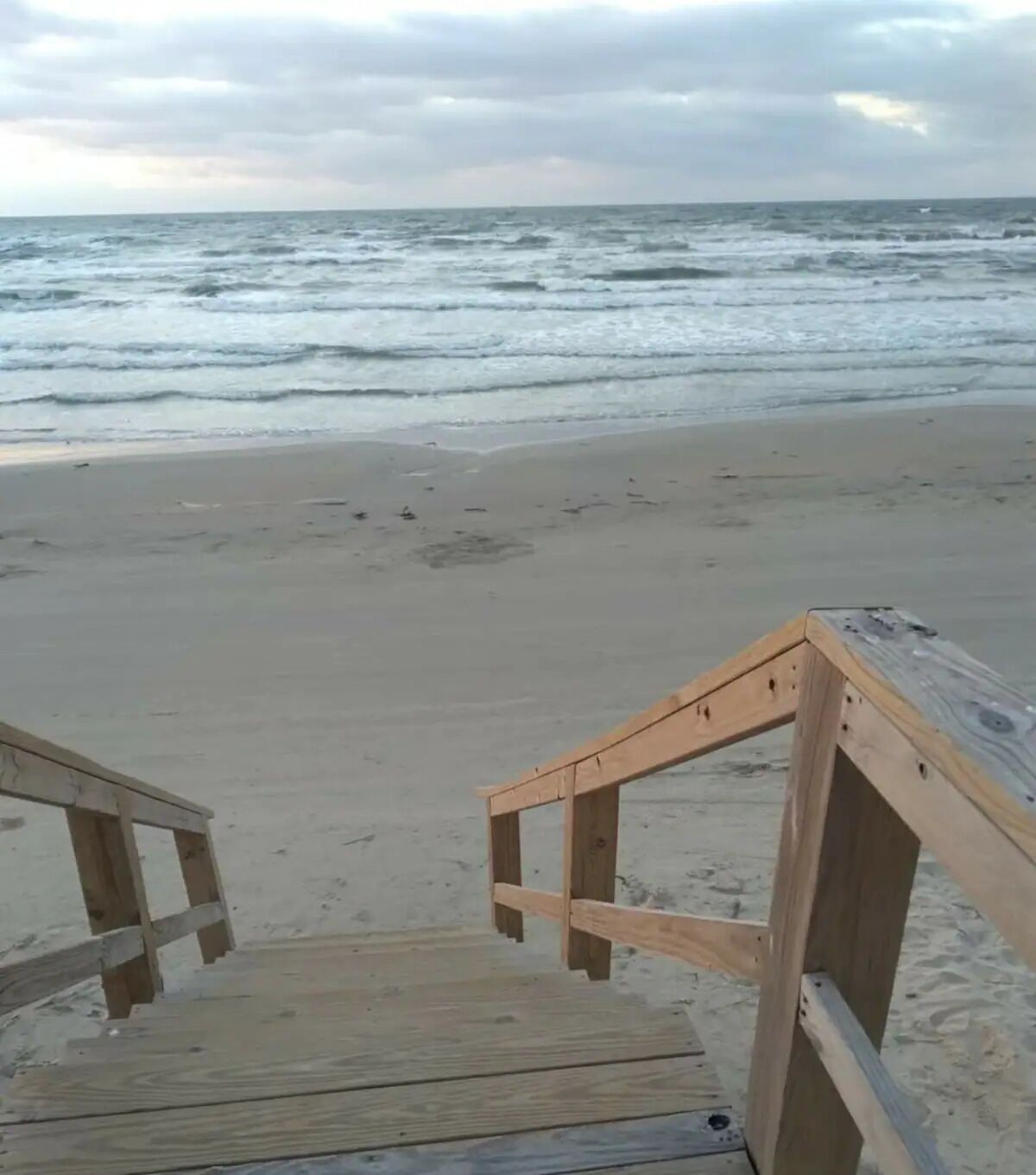 Eine Holztreppe führt zu einem Strand, im Hintergrund sind Wellen zu sehen.