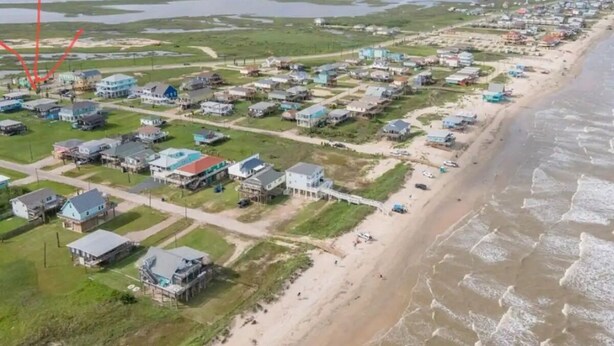 Vista aérea de un pueblo costero con casas a lo largo de la playa, cielo despejado y una masa de agua al fondo.