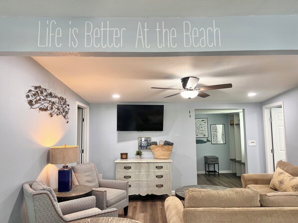 A living room with a ceiling fan, a TV, and a wall sign that reads "Life is Better At the Beach".