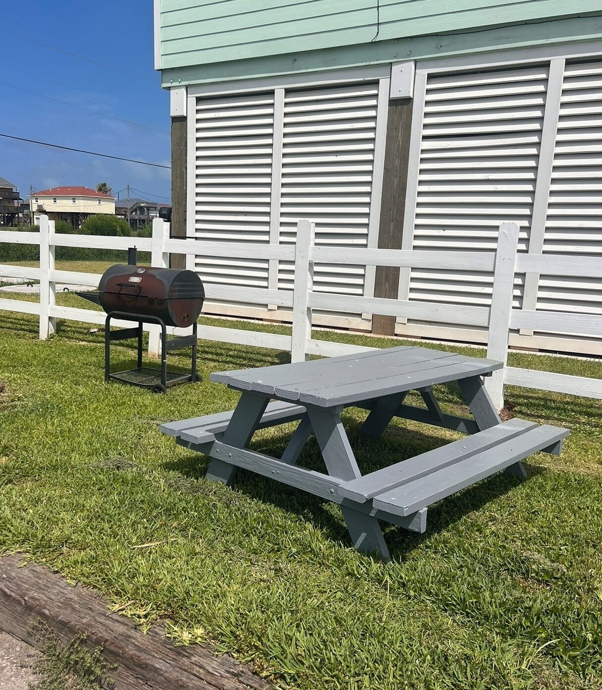 A picnic table and grill set up in a grassy area with a white fence and a building in the background.