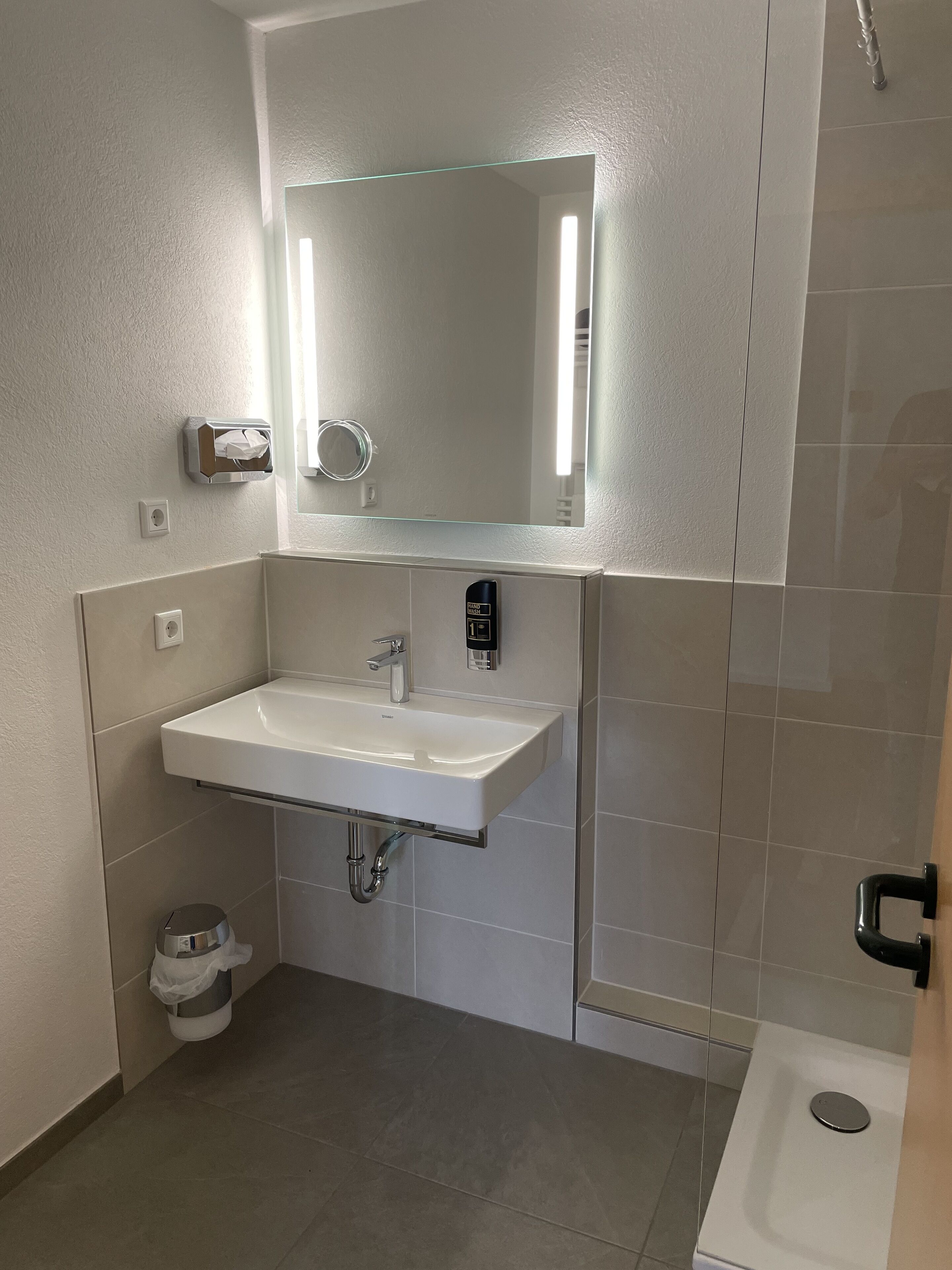 A modern bathroom with a white sink, a mirror, a towel dispenser, and a shower.