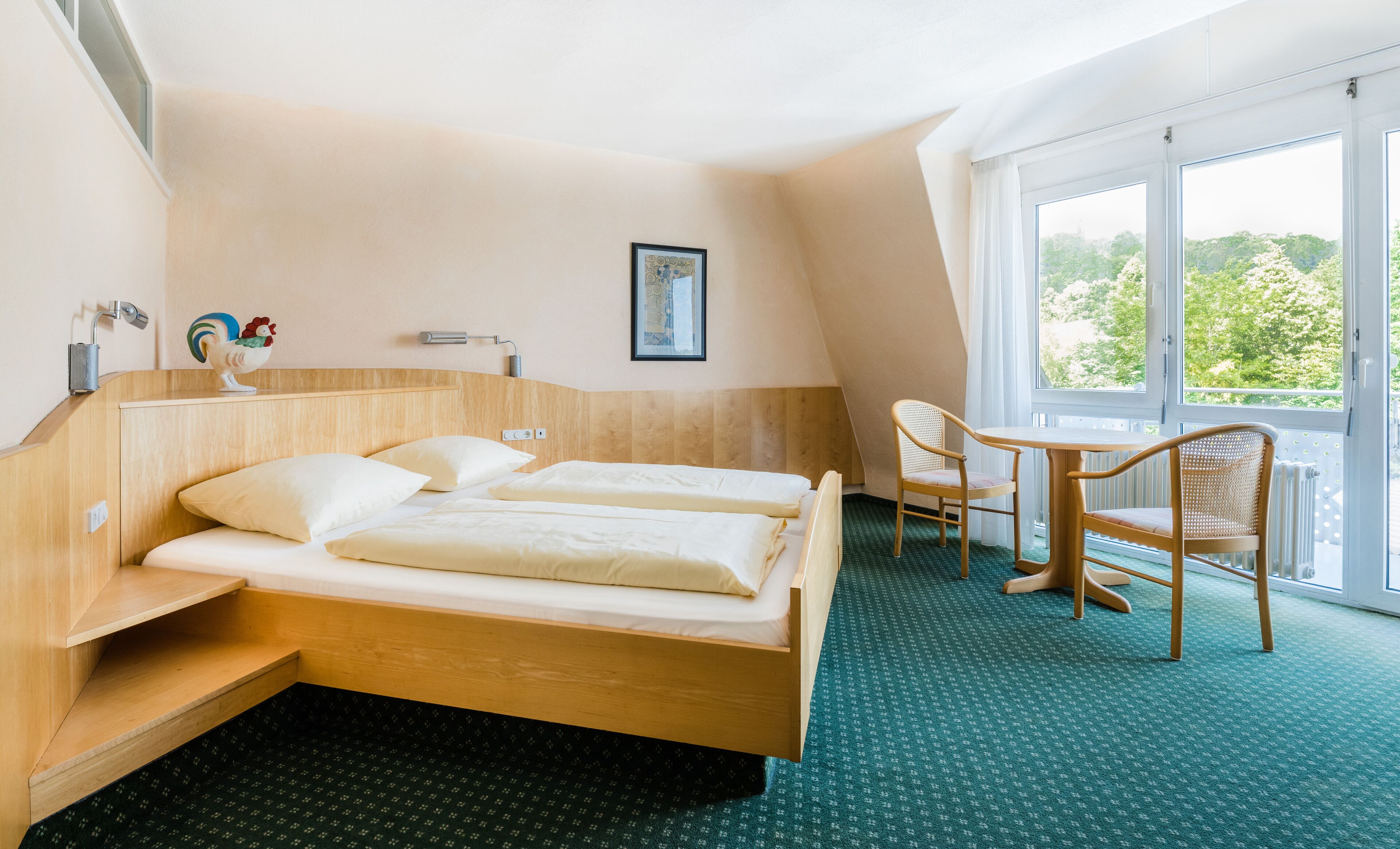 A hotel room with a bed, a chair, a small table, a window with a view of greenery, and a picture on the wall.