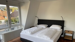 A modern bedroom with a large bed, a wooden floor, a radiator, and a window with a view of houses. - Bikehotel Freiburg (Merzhausen)