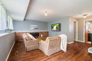 Ein Wohnzimmer mit Kamin aus Ziegelsteinen, zwei beigen Sofas, einem Couchtisch und einem an der Wand montierten Fernseher.