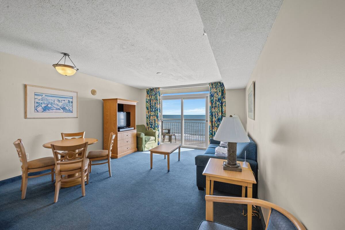 A room with a dining table, chairs, a sofa, a TV, and a balcony with a view.