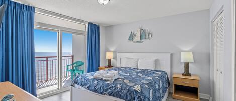 A bedroom with a bed, a nightstand, a desk, and a view of the sea through a balcony.