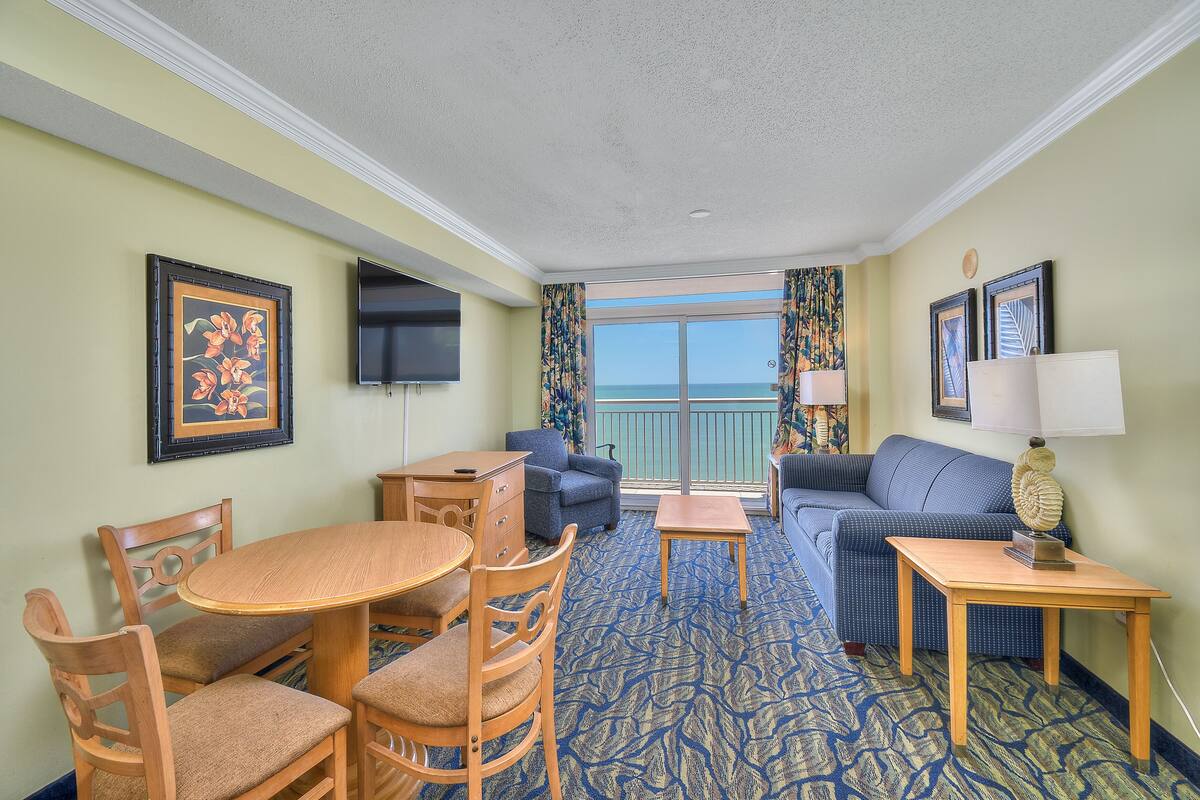 A hotel room with a dining table, chairs, a sofa, a TV, and a balcony with a view of the ocean.