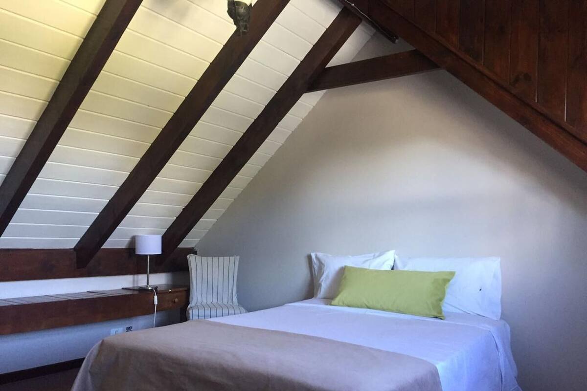 Une chambre avec un plafond en pente, un lit avec des oreillers blancs et verts, une table de chevet avec une lampe et une fenêtre avec des stores.