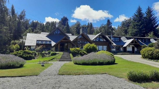 A lodge with a symmetrical design, featuring a central entrance flanked by two wings. A gravel path leads to the entrance, surrounded by manicured lawns and shrubs.
