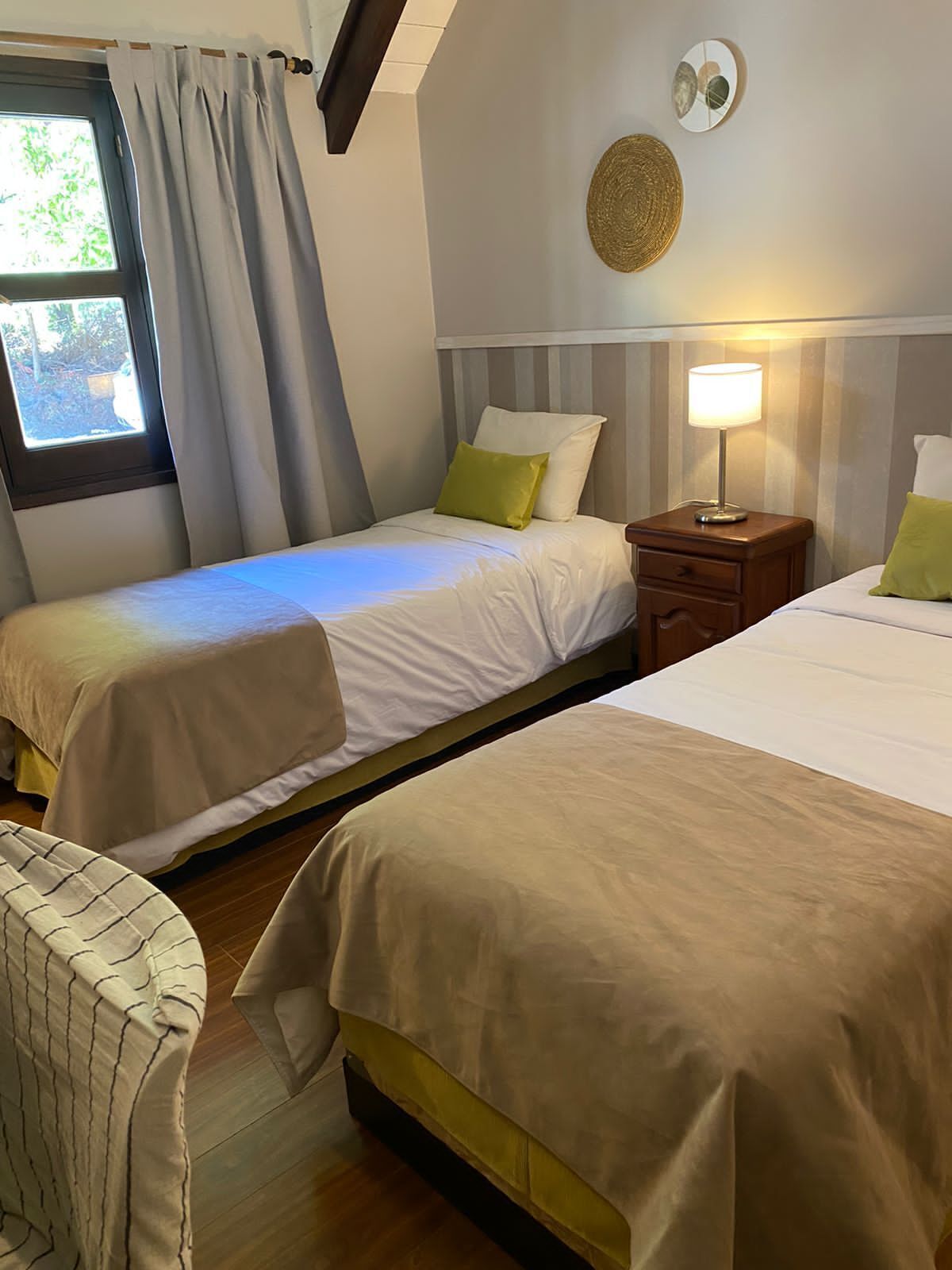 Habitación de hotel con dos camas individuales, una mesita con lámpara, una silla y una ventana con cortinas.