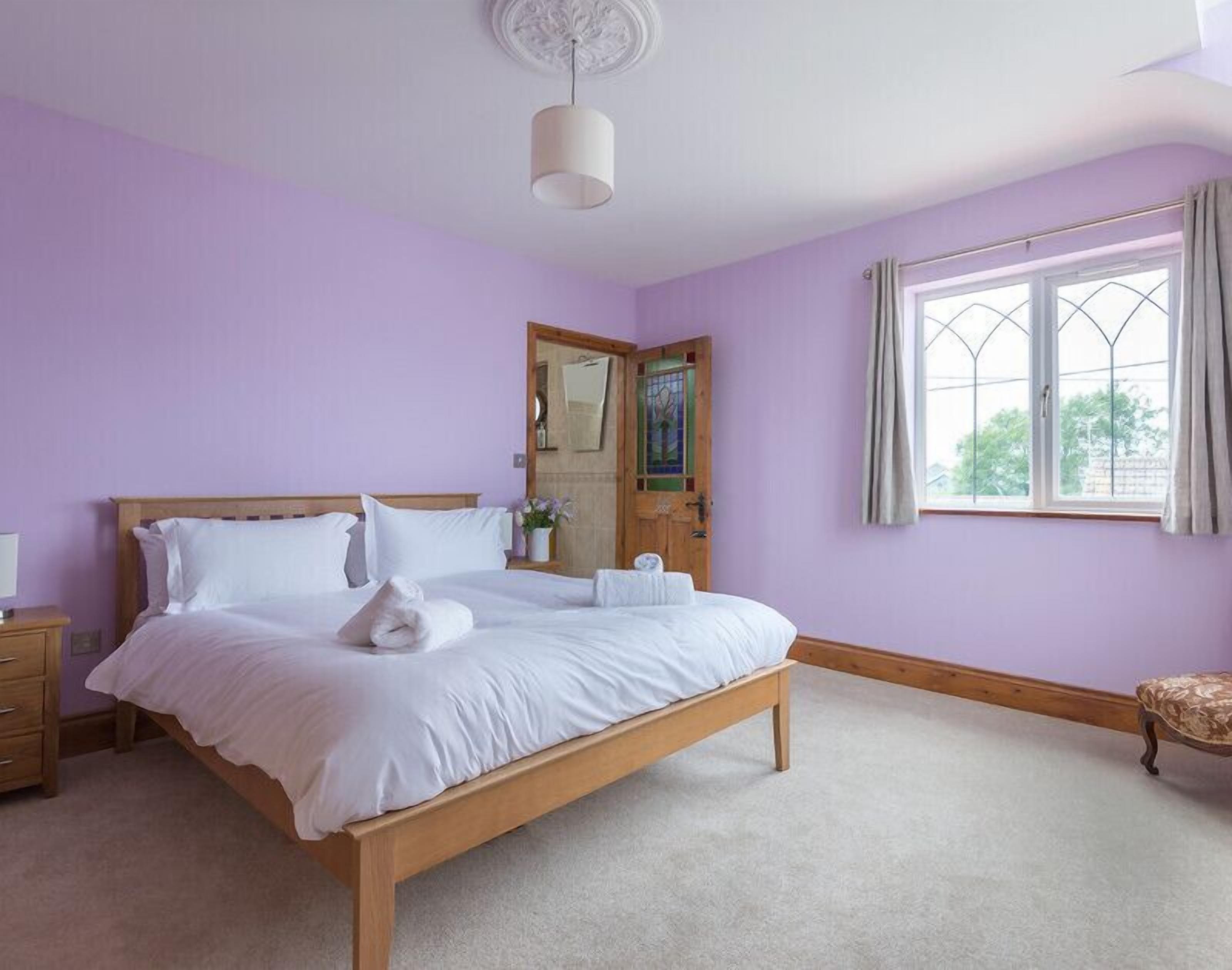 A bedroom with a wooden bed, a bedside table, a nightstand, a window with curtains, and a door with a mirror and artwork.