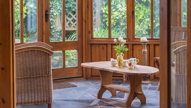 A wooden-paneled room with a round table and wicker chairs, large windows with a view of greenery. - Fairholme (Port Isaac)