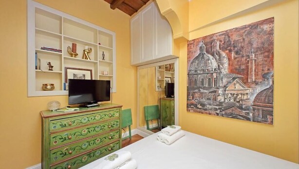 Habitación de hotel con cama, televisión, cómoda verde, un cuadro de un edificio y un espejo.
