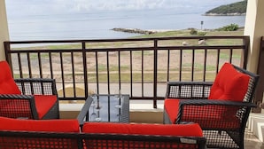 A balcony with wicker furniture and red cushions, overlooking a beach and the sea.