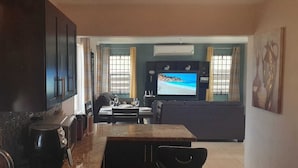 A living room with a TV displaying a beach scene, a sofa, and a dining area with a table and chairs.