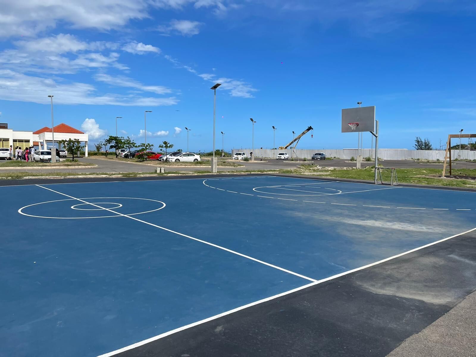 An outdoor basketball court with two hoops, surrounded by a parking area and buildings.