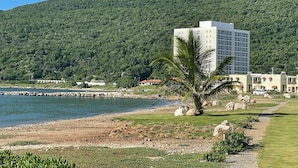 A coastal hotel with a modern building, a palm tree, and a view of the sea and a green hill in the background.