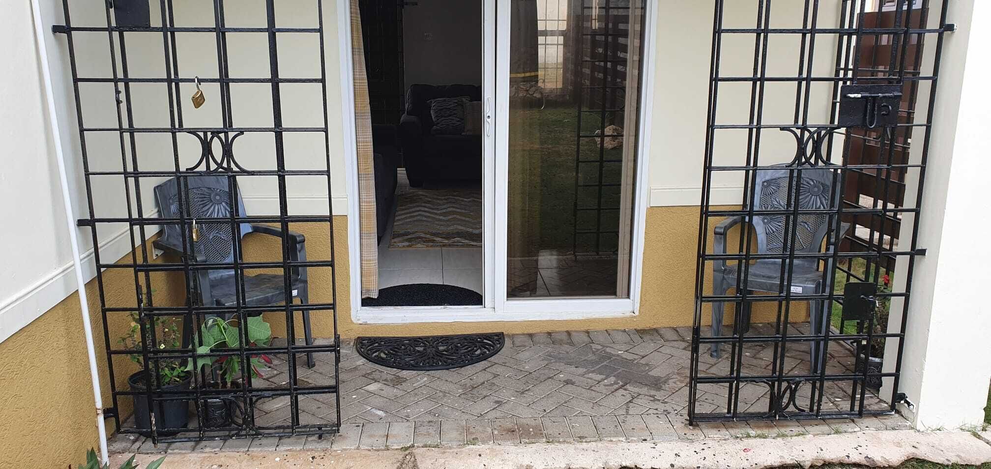 A gated entrance with a sliding glass door, a small outdoor seating area with chairs, and a potted plant.