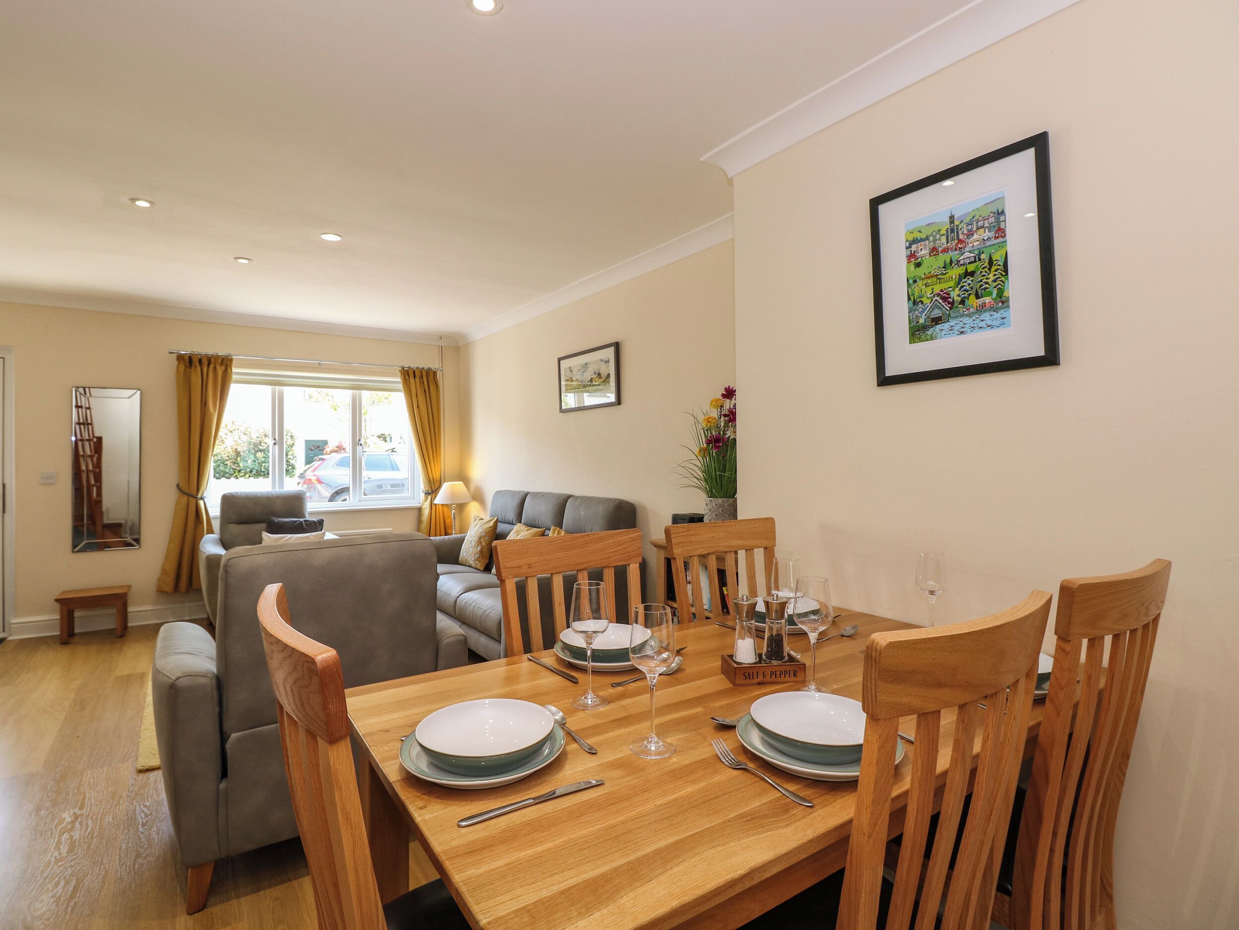 A dining area with a wooden table set for four, surrounded by wooden chairs. There is a sofa and a window with curtains in the background.
