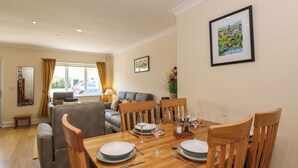 A dining area with a wooden table set for four, surrounded by wooden chairs. There is a sofa and a window with curtains in the background. - Inglewood (Keswick)