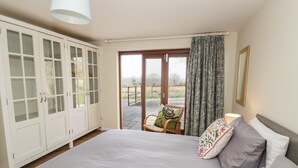 A bedroom with a large bed, a wardrobe, a chair with a cushion, a window with curtains, and a view of the outdoors. - The Wash House (Banbury)