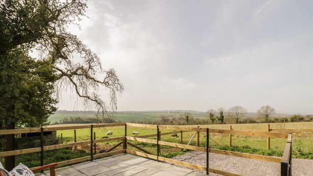 A wooden bench with cushions on a stone patio overlooking a grassy field with a wooden fence. - The Wash House (Banbury)