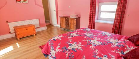 A bedroom with pink walls, a wooden dresser, a radiator, a bed with a floral bedspread, and a window with red curtains.