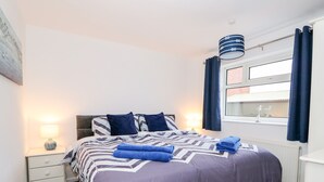 A bedroom with a bed, a nightstand, a lamp, a window with curtains, and a radiator. - Rose Cottage (Skegness)