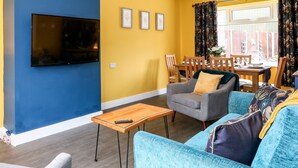 A living room with a TV mounted on a blue wall, a grey armchair, a blue sofa, and a wooden coffee table. - Rose Cottage (Skegness)