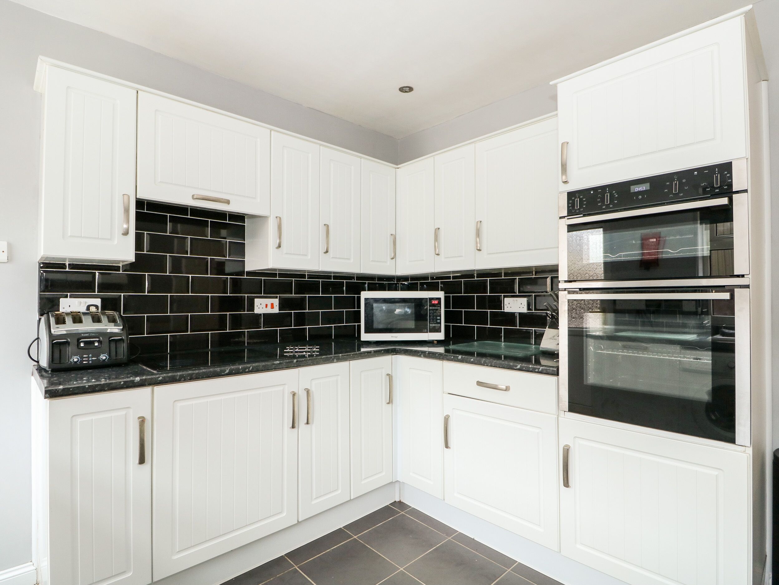 A modern kitchen with white cabinets, black tiled backsplash, and stainless steel appliances including a toaster, microwave, and oven.