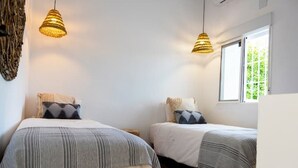 Quarto com duas camas, cada uma com roupa de cama listrada e almofadas geométricas. Há luminárias de fibra trançada acima das camas e uma janela no lado direito.