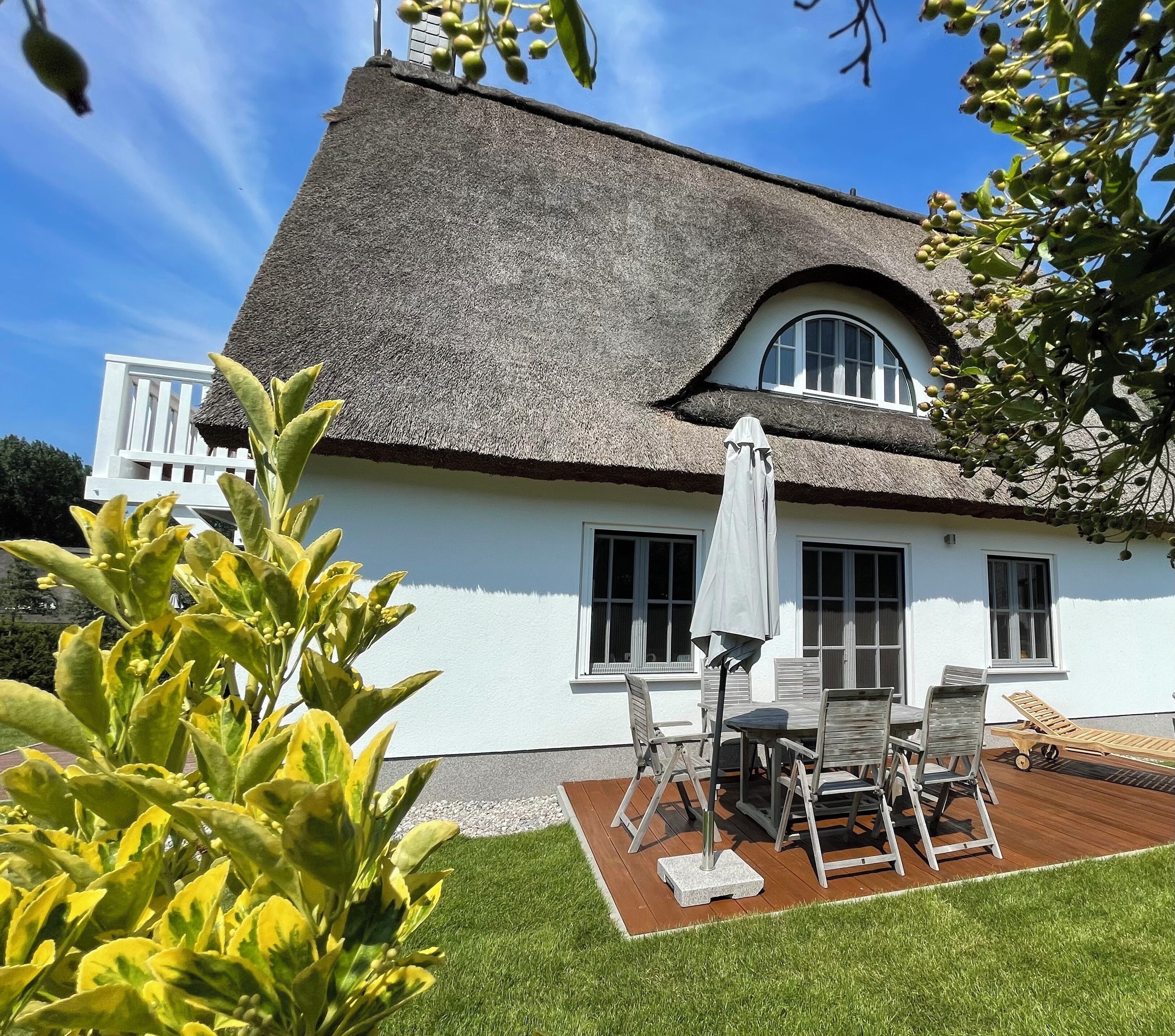 Ein Haus mit einem Strohdach, weißen Außenwänden, einer Terrasse mit Tisch und Stühlen sowie einem kleinen Garten.
