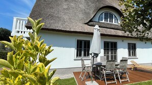 Ein Haus mit einem Strohdach, weißen Außenwänden, einer Terrasse mit Tisch und Stühlen sowie einem kleinen Garten.