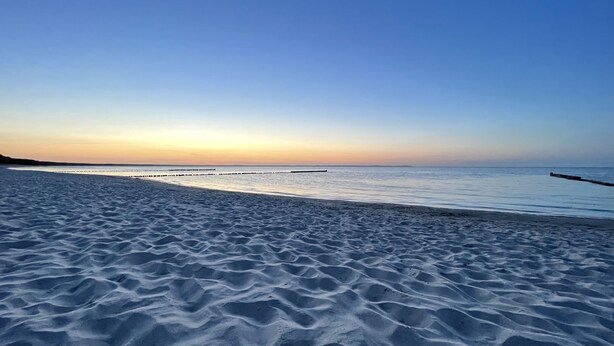 Ein Strand mit einer hölzernen Pier, die sich in ruhiges Wasser erstreckt, umgeben von welligem Sand.