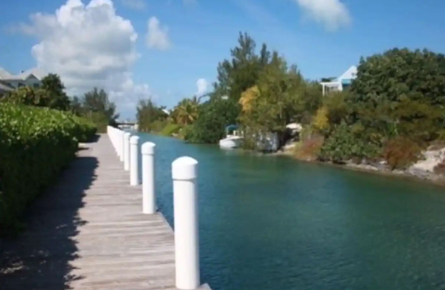 A wooden boardwalk along a calm river, flanked by lush greenery and white bollards.