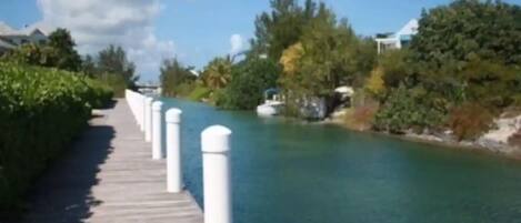 A wooden boardwalk along a calm river, flanked by lush greenery and white bollards.