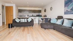 A modern living room with a sofa and armchair, a dining area with a table and chairs, and a kitchen in the background. - Fistral Apres Surf (Newquay)