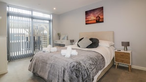 A modern bedroom with a large bed, a nightstand, a bedside table, and a view through a sliding glass door. - Fistral Apres Surf (Newquay)