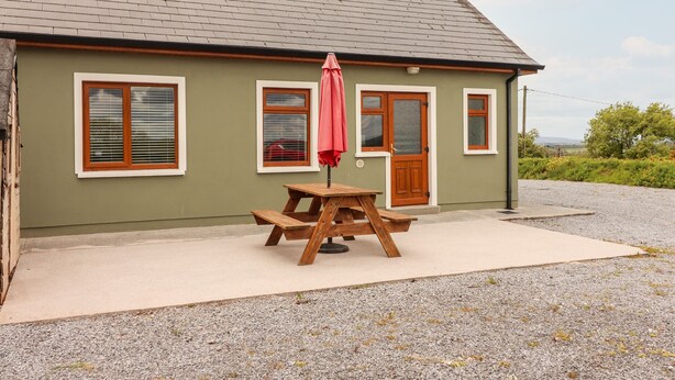 A house with a gravel driveway, a wooden picnic table with a red umbrella, and a door with glass panels. - Daisy's Cottage (Knockreagh)
