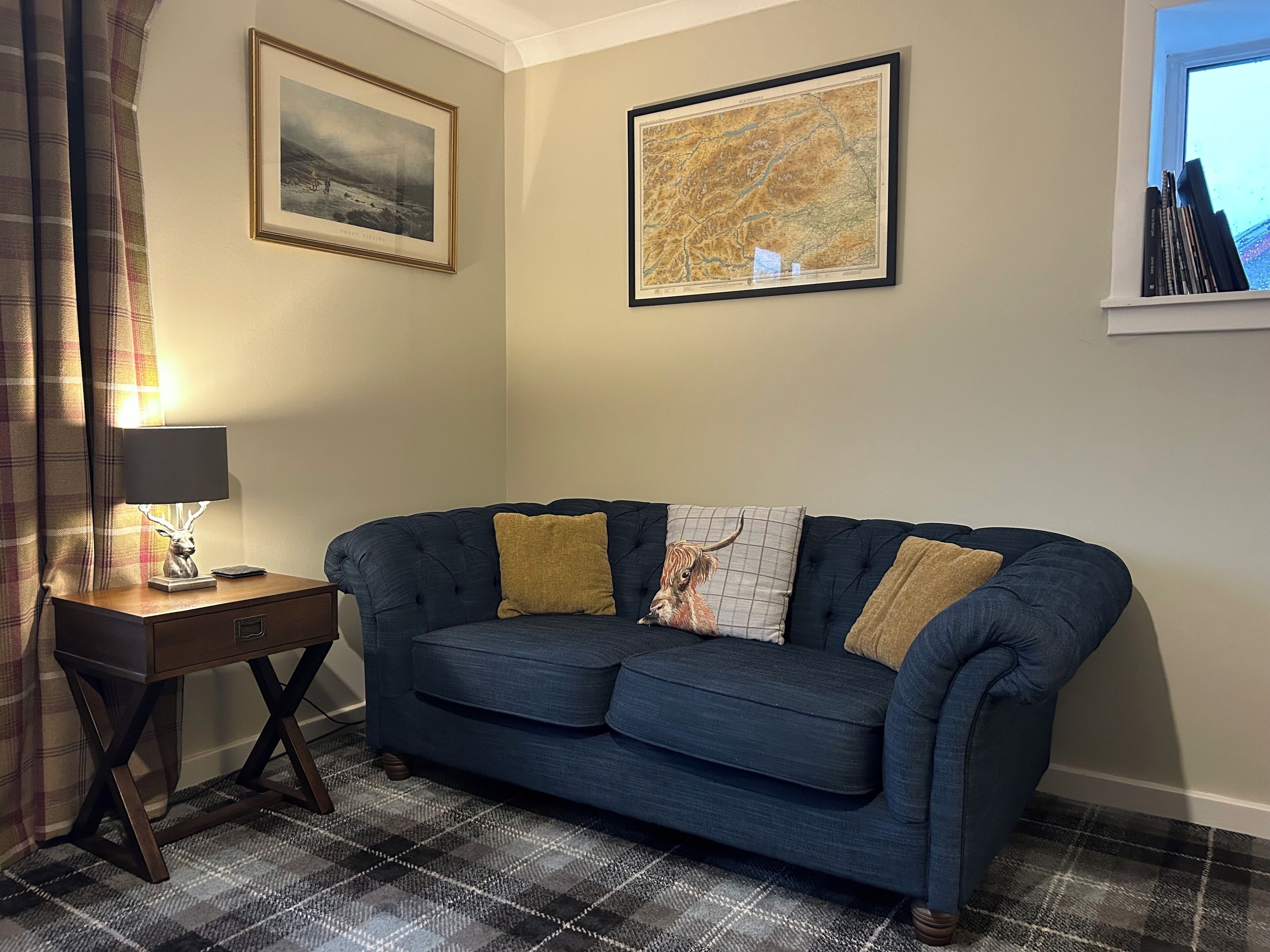 A room with a dark blue sofa, a small wooden side table with a lamp, a plaid-patterned rug, and two framed pictures on the wall.