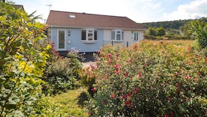 A single-story house with a tiled roof, surrounded by lush greenery and flowering plants. - Tillys Cottage (Weston-super-Mare)
