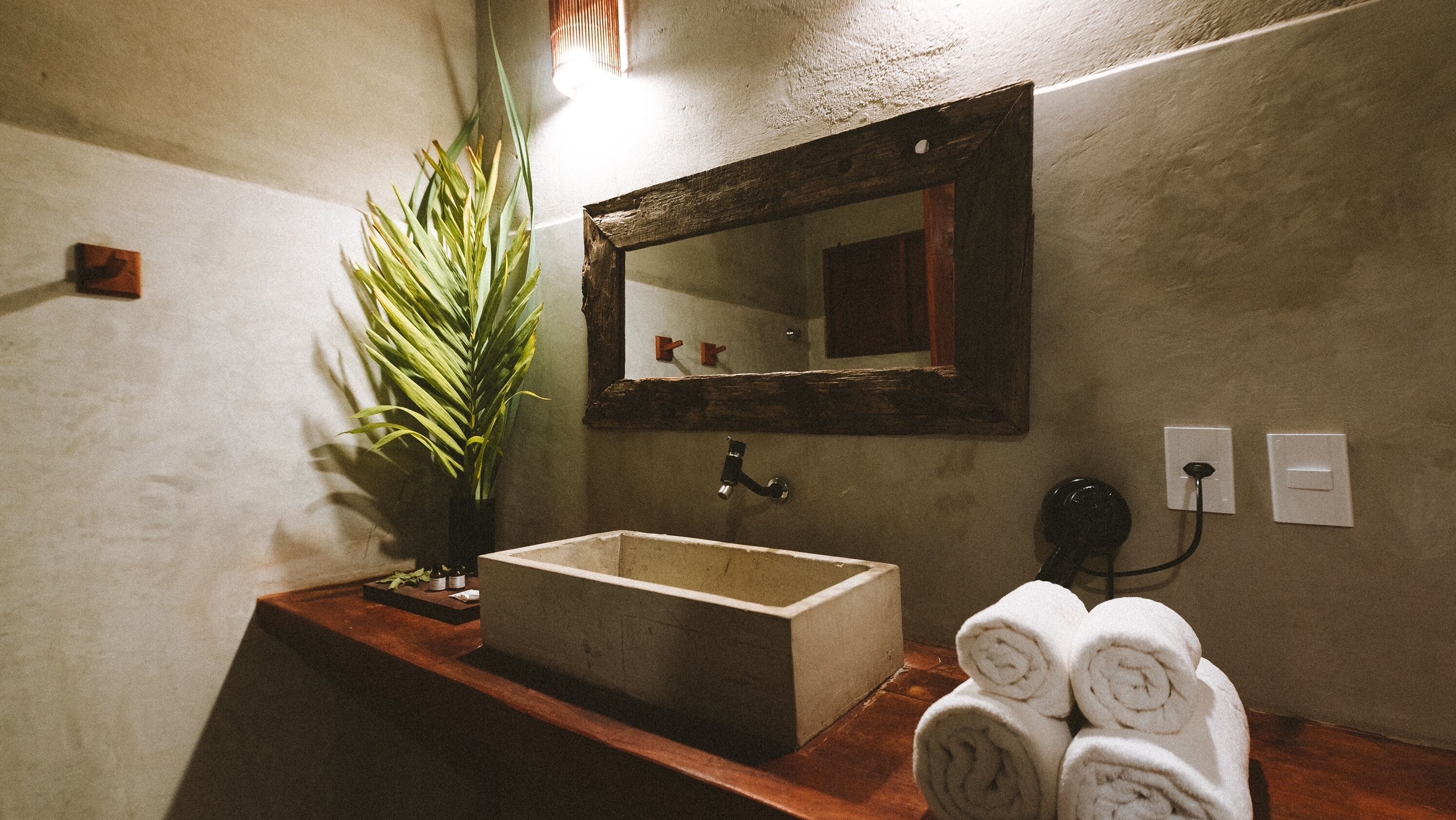 A bathroom with a wooden vanity, a sink, a mirror, a potted plant, and rolled towels.