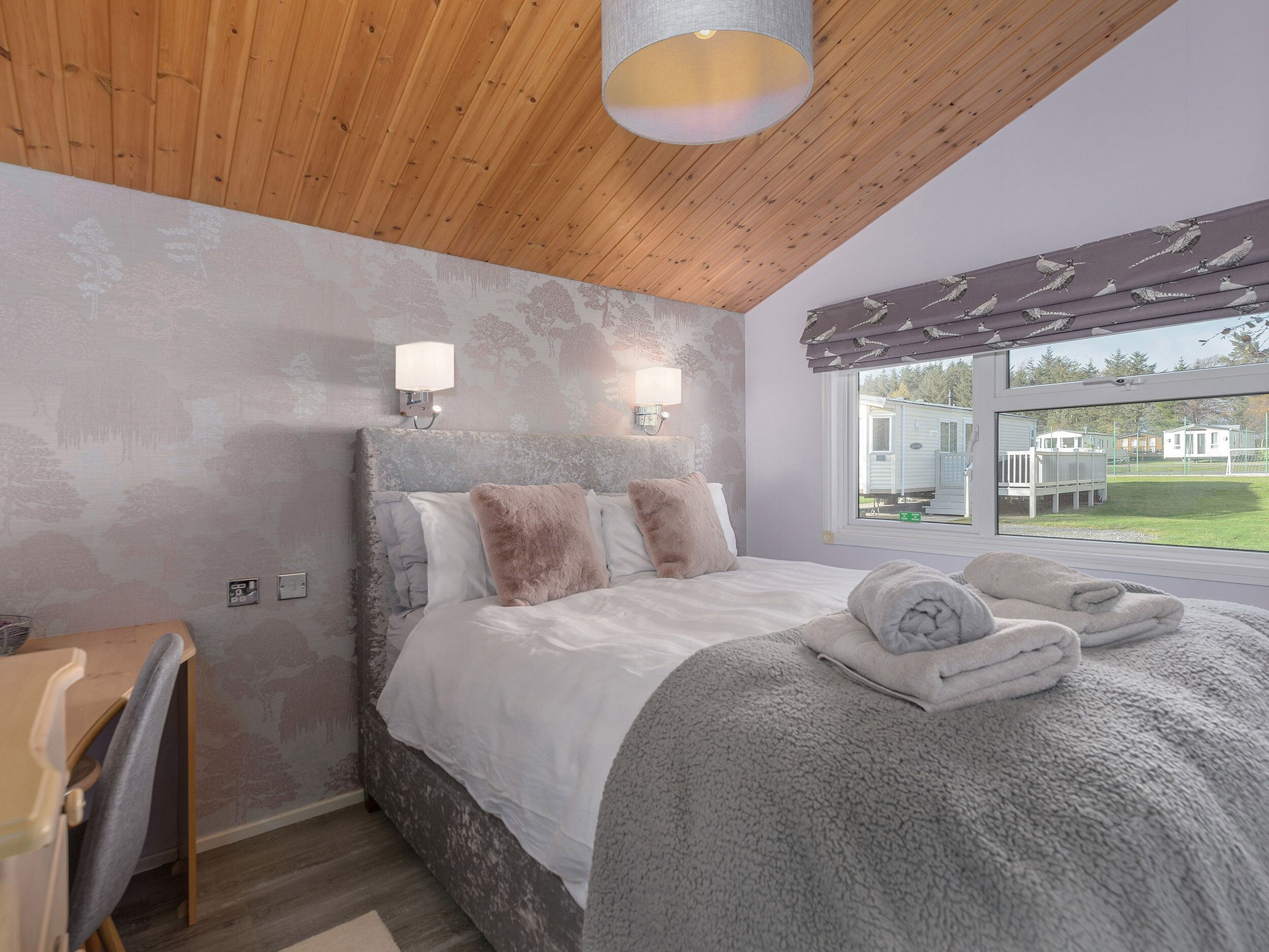 Un dormitorio con techo de madera, una cama con manta gris, dos luces empotradas en la pared y una ventana con vistas a viviendas móviles.